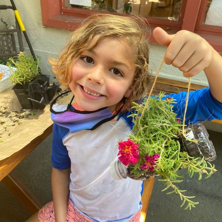 A Boy is holding up a planted and hangable plant towards the camera while smiling widely.