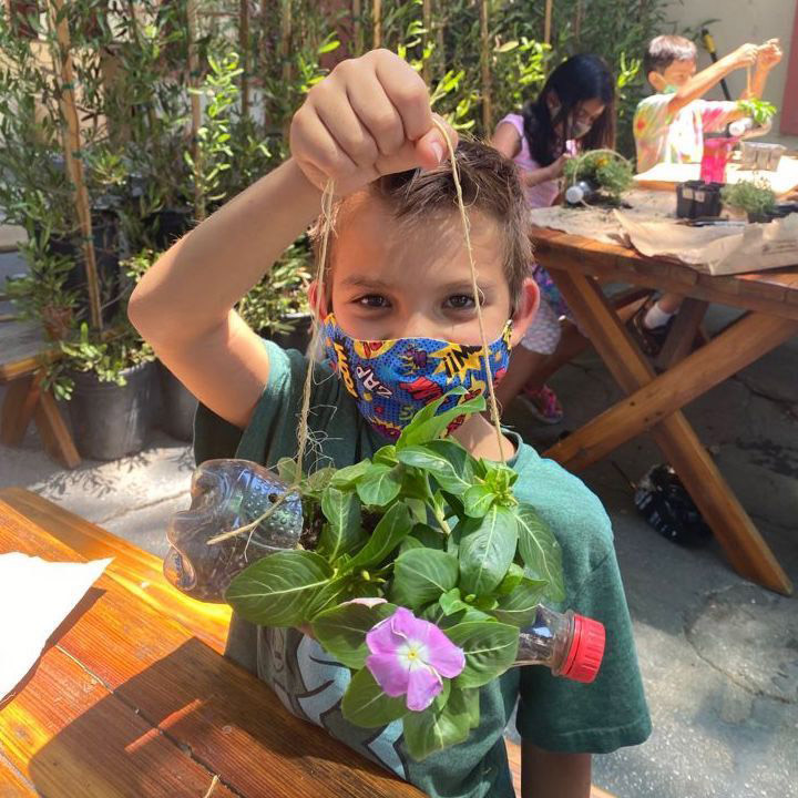 A Boy is holding up a planted and hangable plant towards the camera while smiling widely.