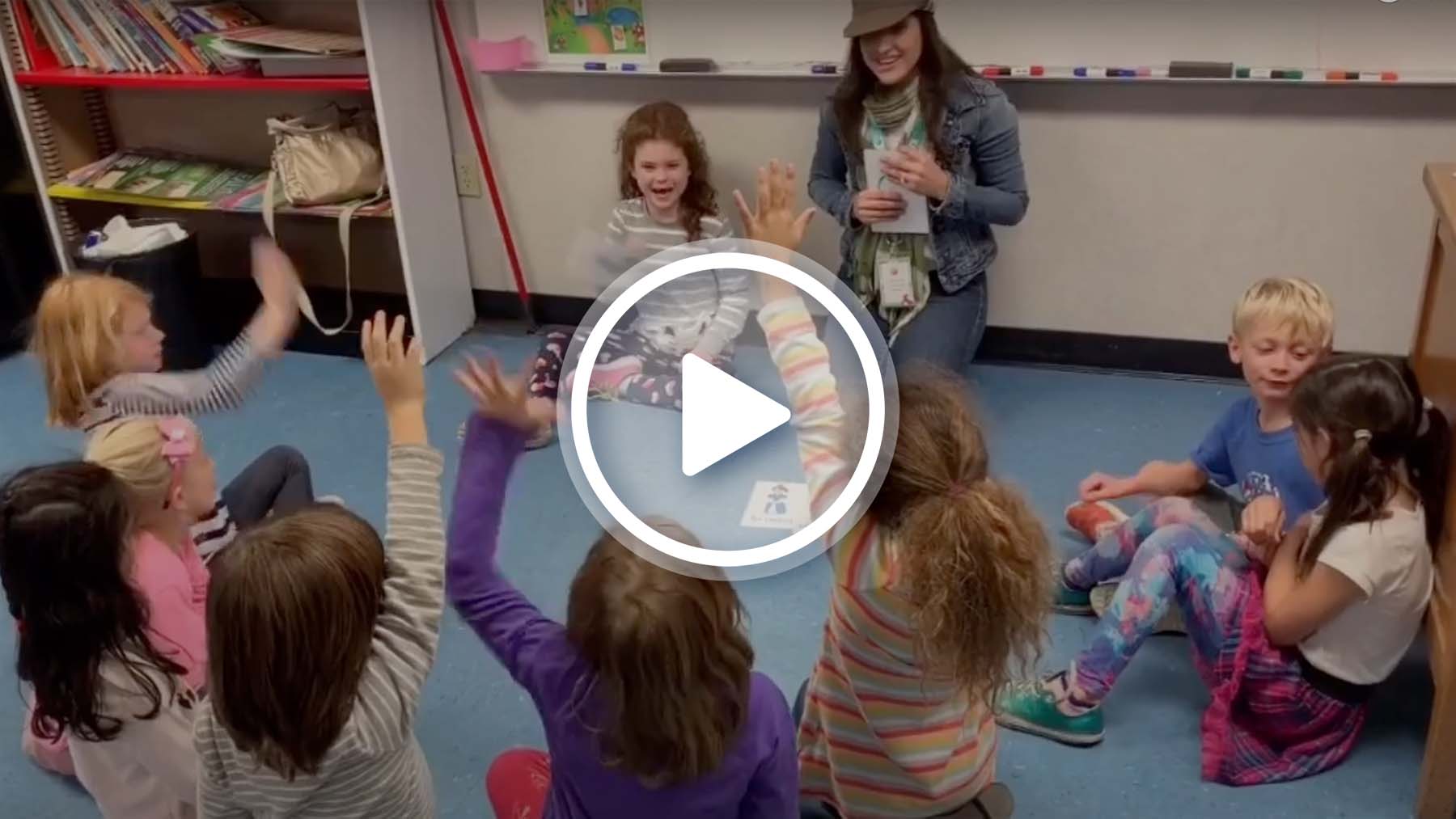 Group of students with teacher sitting on floor about to play a game with Spanish vocabulary cards. One student is next to teacher facing the group.