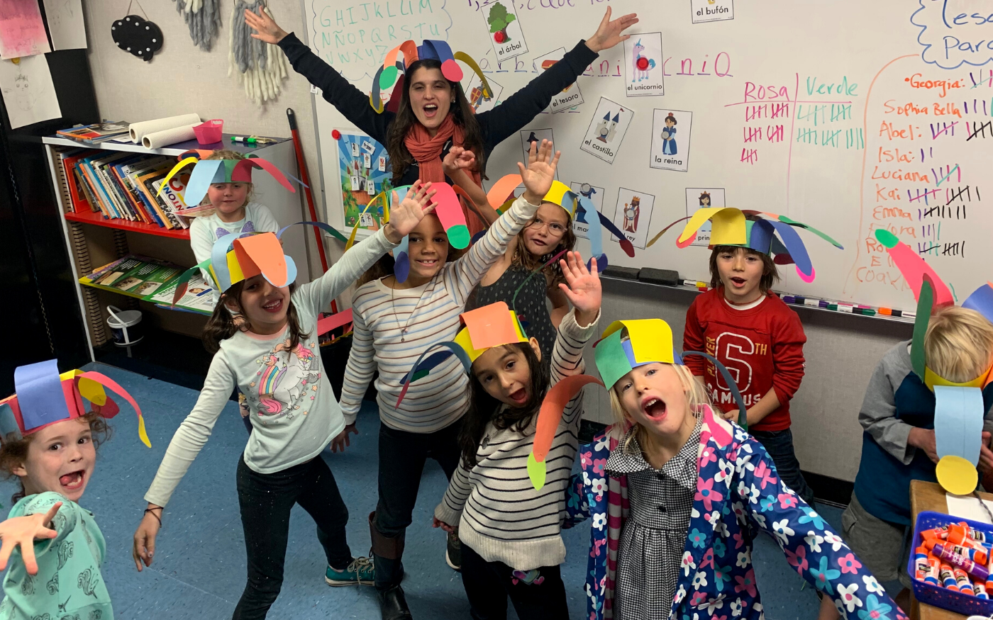 Nine children and their teacher making silly poses for a picture in the classroom wearing hand-crafted paper hats