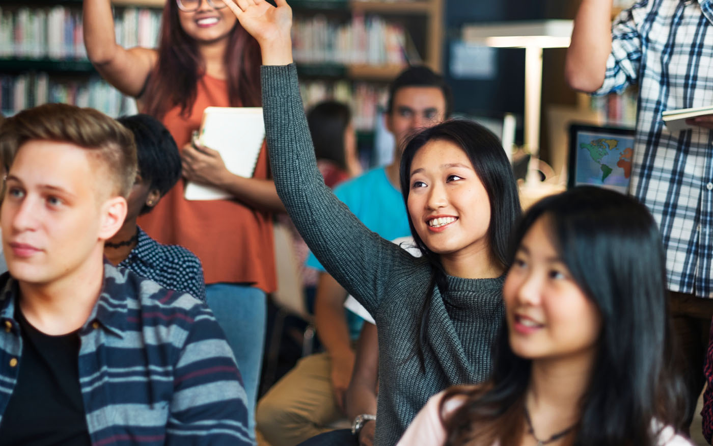 A diverse group of High School students are listening to a lecture and one smiling student is raising her hand.