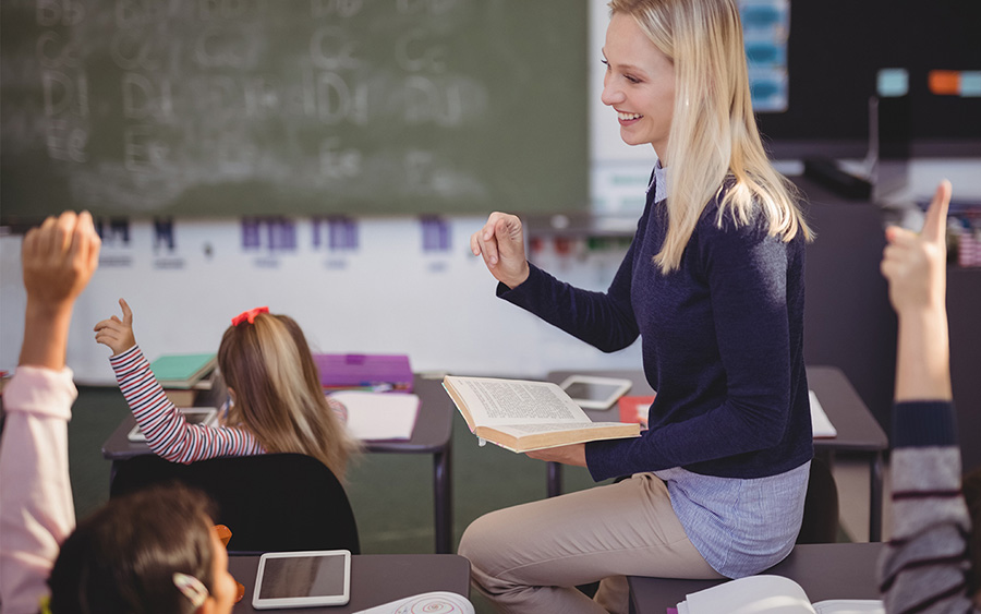 School kids raising their hands at their desks while teacher holding a book open resting in one hand is looking at class and smiling.