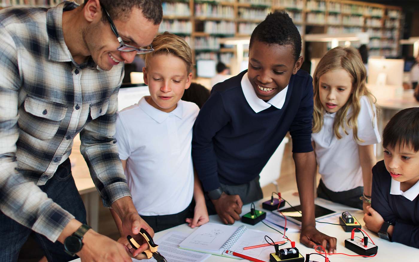 A small, diverse group of elemetary school children are gathered around a teacher at a table demonstrating a physics model.