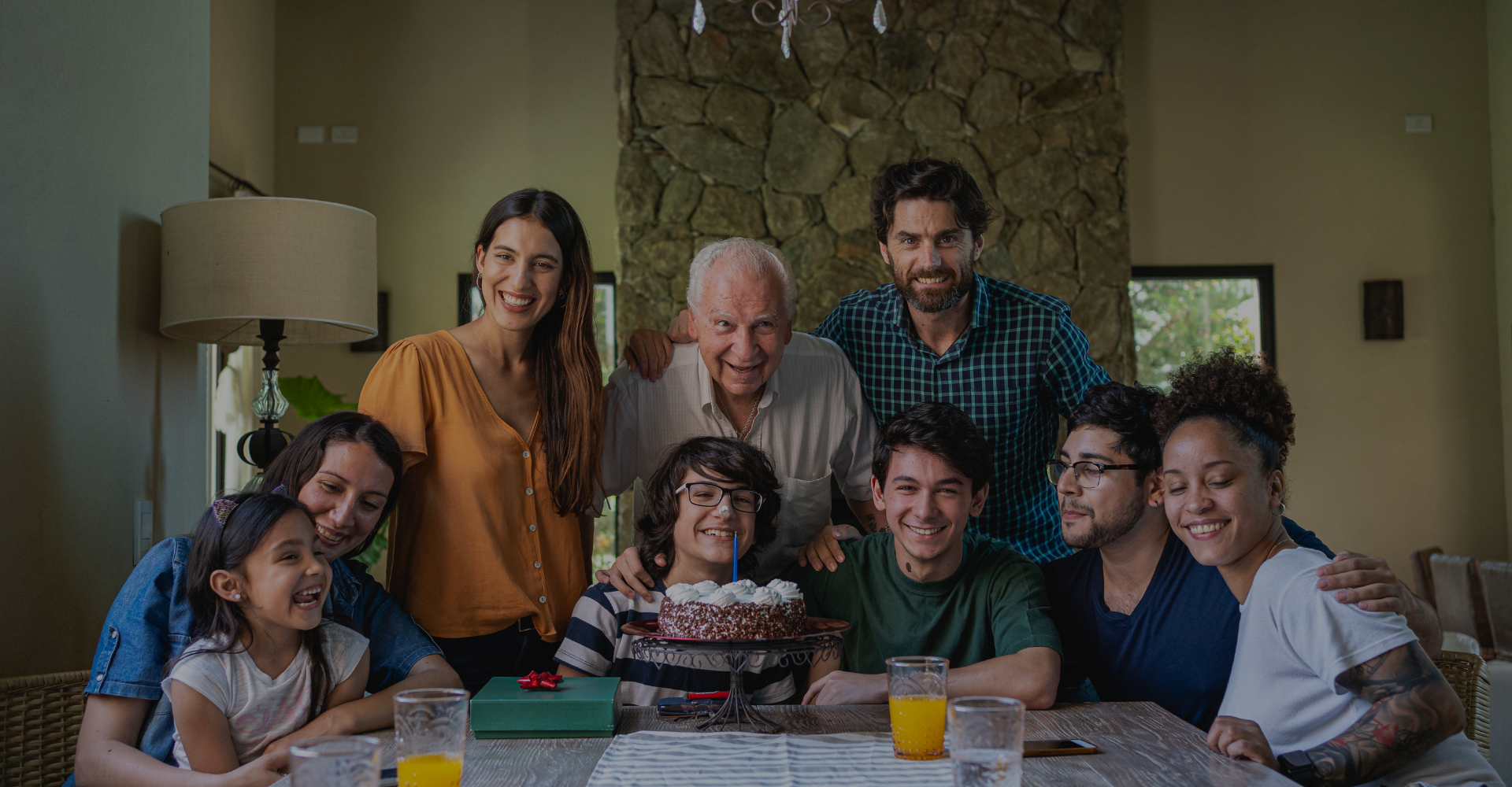 A multi-generational family of 9 celebrating a birthday around the dining table with cake and gifts.