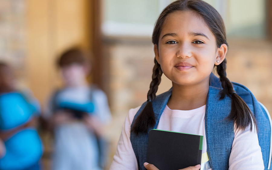 Portrait of smiling face of elementary girl with backpack holding a book.