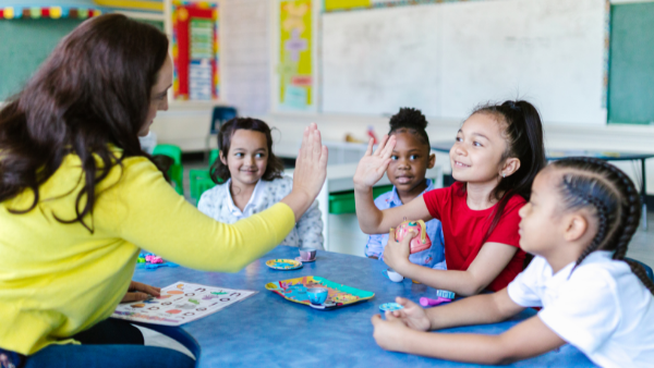 Four children gathered in a semi-circle around their teacher in a classroom; one child is giving their teacher a high-five